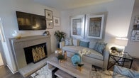 This single-story home in Lewisville, Texas, offers a thoughtfully designed living space with a warm and inviting ambiance. The living room serves as the focal point of the home, showcasing a tile-surround fireplace that adds both character and comfort. A wall-mounted flat-screen television is positioned above framed artwork, creating a curated gallery-style display. The seating area features a neutral-toned sofa accented with chevron and solid teal throw pillows, complemented by a pair of large silver-framed abstract artworks that lend a sophisticated, contemporary feel. A rustic wood coffee table anchors the space, styled with decorative vases in teal and gold tones, fresh florals, and greenery that add a natural, lively touch. The light color palette of whites, grays, and soft blues creates an airy, coastal-inspired atmosphere throughout. A textured area rug adds depth underfoot, while the overall layout maximizes the sense of space. The combination of natural elements, metallic accents, and soft tones gives this Lewisville home a polished yet approachable character.