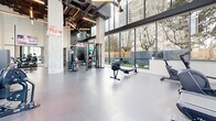 Located at 612 7th Avenue in New Westminster, BC, this contemporary fitness facility — known as The Pulse — offers a generously proportioned, single-floor workout space designed to accommodate a wide variety of training styles. The gym's most striking architectural feature is its double-height ceiling, which creates an open, airy atmosphere and is complemented by exposed concrete columns, industrial-style ductwork, and modern linear lighting fixtures overhead.

Floor-to-ceiling windows line the perimeter walls, flooding the interior with abundant natural light and providing views of the surrounding neighbourhood and greenery. The durable rubberized flooring throughout is both functional and visually cohesive, supporting everything from heavy lifting to dynamic movement.

The equipment layout is thoughtfully organized: a dedicated cardio zone features treadmills, ellipticals, and rowing machines, while a strength training area includes cable machines, a full dumbbell rack with adjustable benches, and multi-functional cable systems. A functional training section offers kettlebells, stability balls, and additional accessories. Large wall-mounted mirrors and a mounted television enhance the space further. An adjacent outdoor green space is visible through the windows, adding to the facility's bright, welcoming ambiance.