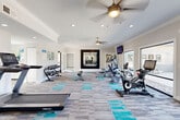 Located at University Station Circle in Charlotte, NC, this single-floor fitness facility offers a thoughtfully designed space for health and wellness. The layout comprises a generously sized exercise room and a flexible open living area, together providing ample room for a full range of fitness activities.

The main exercise room is bright and airy, featuring large windows and glass doors that flood the space with natural light while offering views of the surrounding outdoor area. Ceiling fans and recessed lighting enhance comfort throughout. The room is outfitted with an array of cardio equipment, including treadmills, stationary bikes, and an elliptical, as well as a dedicated free weights station with a full dumbbell rack. Wall-mounted televisions are positioned for convenient viewing during workouts, and a large framed mirror adds depth to the space.

A separate, open-plan room serves as a functional flex area, currently utilized for stretching and group fitness. It features resistance bands, stability balls, medicine balls, and mats neatly organized along the wall. Crown molding and neutral gray tones with teal carpet accents lend a clean, modern aesthetic throughout. An additional entry point connects directly to the outdoors, reinforcing the facility's bright and welcoming atmosphere.