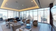 This elegant single-floor space is designed to impress, combining contemporary architecture with sweeping urban panoramas. The defining feature of the room is its expansive floor-to-ceiling glass curtain wall, which wraps around the space and frames commanding views of a dense city skyline. Natural light floods the interior throughout the day, creating a bright and open atmosphere.

The ceiling design is a standout architectural element, featuring a recessed tray configuration with integrated ambient uplighting that adds warmth and visual depth to the room. Recessed spotlights are evenly distributed across the ceiling, ensuring well-balanced illumination during evening hours.

The lounge area is thoughtfully arranged with a collection of neutral-toned upholstered armchairs and sofas, complemented by minimalist glass-and-metal coffee tables. The seating arrangement encourages both intimate conversation and open gathering. Flooring transitions between carpet in the seating zones and hard tile near the window perimeter, adding subtle texture and definition to the layout.

Overall, the space exudes a polished, professional ambiance suited for high-end hospitality, corporate lounging, or executive entertaining, with its refined finishes and dramatic cityscape backdrop serving as its most memorable attributes.