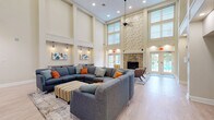 Located at 1100 Belmont Blvd in Monroeville, PA, this two-floor amenity facility offers a generous and well-rounded layout designed for both recreation and community gathering. The main floor commands the majority of the space, while the upper level provides an additional area that overlooks the floors below via open railings, contributing to a sense of volume and grandeur throughout.

The central living room serves as the heart of the facility, boasting soaring double-height ceilings, light hardwood flooring, and elegant wall niches illuminated by pendant lighting and accented with contemporary artwork. Comfortable sectional seating and a wall-mounted television create a welcoming communal atmosphere. Large entry doors invite natural light, enhancing the bright, airy ambiance.

Two dedicated exercise rooms cater to active residents. One is equipped with treadmills, ellipticals, exercise balls, and yoga mats, benefiting from high ceilings and large windows with outdoor views. The second doubles as a racquetball and basketball court, featuring polished hardwood floors with court markings, white paneled walls, and a regulation basketball hoop.

The upper level offers an open mezzanine-style area with carpeted floors, decorative columns, white railings, and track lighting. An office and additional unclassified rooms provide functional support spaces. Six carpeted hallways with neutral tones and recessed lighting connect all areas seamlessly throughout the facility.