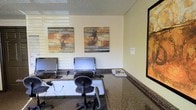 This single-floor space functions as a dedicated business center, thoughtfully arranged to support focused work and productivity. The room features a continuous granite-style countertop running along the walls, providing ample workspace for multiple users simultaneously. Two computer monitors are mounted on adjustable stands atop the counter, accompanied by ergonomic task chairs that allow for comfortable extended use. The countertop's dark stone finish adds a professional and polished aesthetic to the room.

The walls are painted in a neutral, warm cream tone that contributes to a calm and focused atmosphere. Three framed abstract artworks are displayed throughout the space—two positioned above the workstation counter and one mounted on the adjacent wall—each featuring earthy tones of rust, brown, and gold that lend a cohesive and inviting visual character to the room. A textured ceiling adds subtle architectural detail overhead.

Natural light filters into the space through a window fitted with horizontal blinds, offering adjustable brightness to suit different working needs. A small selection of reading materials rests on the counter, adding a practical, welcoming touch to this well-equipped, functional workspace.