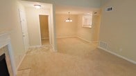 This single-floor residence offers a clean, functional layout with an open-concept living and dining area that flows seamlessly together. Plush, neutral-toned wall-to-wall carpeting extends throughout the main living spaces, providing a warm and comfortable feel underfoot. The walls are finished in a soft beige tone, contributing to a bright and welcoming ambiance.

The dining area is highlighted by a classic chandelier that serves as a subtle focal point, adding a touch of elegance to the otherwise understated space. A pass-through window connects the dining area to the kitchen, promoting an open feel while maintaining a defined cooking zone. The kitchen appears to feature overhead cabinetry and receives natural light from a window, keeping the space feeling airy.

A doorway visible from the main living area leads to an additional room, suggesting a versatile floor plan suited for various living arrangements. Ceiling-mounted light fixtures, HVAC vents, and white baseboards throughout reflect practical, well-considered finishes. Overall, the space presents a straightforward, move-in-ready environment with a neutral palette that allows for easy personalization.