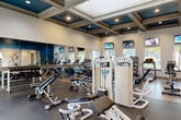 This fitness center offers a generously sized, single-floor exercise space designed to support a full range of workout routines. The room's most striking architectural feature is its elegant coffered ceiling, finished in deep navy blue and cream tones with recessed lighting, lending the space a refined yet energizing ambiance. A large skylight allows natural light to pour in from above, complementing the multiple windows along the walls that provide views of the surrounding outdoor greenery.

The exercise room is outfitted with a comprehensive selection of equipment. The cardio zone features treadmills, elliptical trainers, stationary bikes, and stair climbers from brands including Cybex and LeMond. The strength-training area includes cable machines, adjustable benches, free weight racks, and a variety of resistance machines. Full-length mirrors line one wall, visually expanding the space and aiding users during workouts.

Flooring transitions between rubberized mat sections in the strength area and wood-look flooring in the cardio zone, offering both safety and style. Multiple flat-screen TVs are mounted throughout, providing entertainment options during workouts. A built-in counter with granite surfaces near the entrance adds a functional touch. Ceiling fans contribute to comfortable air circulation, completing this thoughtfully designed fitness facility.
