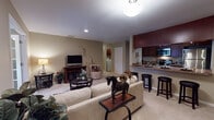 Located at 107 Bent Tree Lane in Schaumburg, IL, this single-floor residence offers a well-designed, open-concept layout with distinct functional areas flowing seamlessly into one another. The living room serves as the centerpiece of the home, featuring plush carpeting, recessed lighting, and a media console with an electric fireplace beneath a wall-mounted television. Neutral warm-toned walls provide a versatile backdrop throughout the space. An adjacent den, accessible through French-style doors, offers natural light through a window dressed with layered curtains and is well-suited as a home office or secondary sitting room, complete with a ceiling fan.

The galley-style kitchen is efficiently organized with rich dark wood cabinetry, granite-look countertops, stainless steel appliances including a built-in microwave and refrigerator, and a double sink. A breakfast bar with bar stools connects the kitchen to the open living area.

The primary bedroom features a bold red accent wall, carpeted floors, natural light from multiple windows, and ample space for furnishings. Two bathrooms are present—one with a tub-shower combination and one with a single vanity—both outfitted with dark wood cabinetry and clean white fixtures. An in-unit laundry area is also visible, adding everyday convenience.