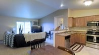 This single-floor residence offers a practical and well-connected open-concept layout, seamlessly integrating the kitchen, dining, and living areas into one cohesive space. The vaulted ceiling adds a sense of height and openness throughout the main living area, enhancing the overall sense of space.

The kitchen features warm wood-toned cabinetry, granite-style countertops, a stainless steel dishwasher, and a double sink. A raised breakfast bar peninsula separates the kitchen from the dining area, providing additional counter space and a casual dining option.

The dining area accommodates a traditional wooden table and chairs, positioned conveniently between the kitchen and the living room. The living room is bright and inviting, benefiting from a large sliding glass door that allows generous natural light to fill the space. A wall-mounted air conditioning unit provides climate control, and the carpeted flooring throughout the main living areas adds warmth and comfort.

The neutral gray and beige tones on the walls and flooring create a clean, understated aesthetic throughout. Overall, this space offers a functional and comfortable layout well-suited for everyday living.