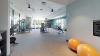 Located at 9235 Senator Royall Dr. in Charlotte, NC, this contemporary fitness center is thoughtfully designed to support a wide range of workout routines. The single-floor facility features an open, spacious layout with neutral-toned carpet flooring and a clean, modern aesthetic enhanced by soft recessed lighting and ceiling fans that keep the environment comfortable during workouts.

The cardio zone is well-equipped with a row of treadmills and elliptical machines positioned along large windows, allowing natural light to pour in and offering views of the surrounding community. A bold, sky-inspired mural adds a vibrant, energizing focal point to one end of the room.

The strength training area includes a variety of cable machines, weight benches, and a multi-functional rack system. Free weight options are neatly organized on a dedicated rack, including colorful kettlebells and medicine balls of varying weights, catering to users of all fitness levels.

A separate stretching and functional training area provides open floor space with stability balls and storage cubbies for personal items. Full-length mirrors line the walls, enhancing the sense of space and allowing users to monitor their form. Glass-panel doors open to an adjacent outdoor area, further connecting the interior to the surrounding environment. The overall ambiance is bright, clean, and motivating.