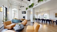Located at 8181 El Mundo St in Houston, TX, this single-floor community clubhouse offers a thoughtfully designed gathering space with a bright, open layout. Soaring ceilings and expansive floor-to-ceiling windows—including striking arched frames—flood the interior with natural light and offer views of the surrounding landscaped grounds and pool area. The main lounge area features a dramatic fireplace, wall-mounted TV, and an eclectic mix of curved seating in warm earth tones, complemented by hanging greenery along the upper columns that adds a fresh, organic touch. Modern ceiling fans and sleek wall sconces provide both function and ambiance throughout. A large conference-style dining area accommodates group gatherings around a substantial wooden table with upholstered seating, illuminated by an elegant brass chain chandelier with pendant lights. Arched windows in this zone frame lush garden views and invite in generous daylight. The kitchenette features dark cabinetry, a textured tile backsplash, granite countertops, and a curved bar with barstool seating—ideal for casual use. The entry lounge offers a welcoming transition space with caramel leather seating and views to the exterior. Warm hardwood-style flooring unifies the space throughout, creating a cohesive and inviting atmosphere.