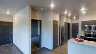 This single-floor residence offers a clean, contemporary design with a cohesive aesthetic carried throughout. The interiors are finished with rich, dark wood-look plank flooring that contrasts smartly against neutral gray walls and crisp white ceilings, creating a modern and polished look. Dark-trimmed doors and baseboards add a bold architectural accent that ties the space together.

The open living area is well-lit by natural light streaming through slatted blinds, and recessed ceiling lights provide consistent illumination throughout. A dedicated laundry closet with side-by-side washer and dryer units offers in-unit convenience, neatly tucked behind bifold doors.

The kitchen is equipped with stainless steel refrigerator and features granite countertops, lending a refined touch to the functional space. Multiple doorways lead to additional rooms, including carpeted spaces that offer a softer finish underfoot, ideal for bedrooms or private areas.

The hallway corridor efficiently connects the various rooms, with recessed lighting guiding the way. The layout is practical and thoughtfully organized, making excellent use of the available footprint. Overall, this residence combines modern finishes, functional amenities, and a well-structured floor plan to deliver a comfortable and stylish living environment.