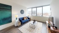 This well-appointed one-bedroom residence combines modern design with practical living across a single thoughtfully arranged floor. The open-concept living area is anchored by expansive floor-to-ceiling windows that frame sweeping urban skyline views, flooding the space with natural light and creating a seamless connection between interior comfort and the city beyond. The living room offers generous space for lounging and entertaining, with wood-look plank flooring that flows throughout the unit.

The kitchen is a standout feature, showcasing dark cabinetry that extends to the ceiling, stainless steel appliances, a built-in microwave, and a large peninsula with a white quartz-style countertop and bar seating — ideal for casual dining or hosting. Pendant lighting adds a refined touch overhead.

The bedroom is a tranquil retreat, benefiting from its own large window with city views. Neutral tones and clean lines give the room a calm, restful ambiance. The full bathroom features dark vanity cabinetry with ample storage, a full-width mirror with integrated lighting, a soaking tub, and warm hardwood-style flooring.

A dedicated laundry closet houses a stacked washer and dryer with additional shelving for storage, adding a layer of everyday convenience to this polished urban residence.