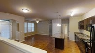 This single-floor space is designed with an open-concept layout that connects the kitchen and living areas into one cohesive, flowing environment. The rich hardwood flooring extends throughout, lending warmth and continuity across the entire space. A standout feature is the kitchen island, topped with a granite-style surface and set on a dark base, offering both functional workspace and a natural visual anchor between the kitchen and living zones.

Overhead lighting is thoughtfully layered: pendant lights hang above the island, a ceiling fan with a light fixture serves the living area, and flush-mount and track lighting provide additional illumination throughout. The neutral-toned walls complement the warm flooring and create a clean, versatile backdrop.

Natural light enters through a set of slatted blinds on one wall, brightening the living area without overwhelming it. Doorways visible in the background suggest access to additional rooms or corridors, providing a sense of depth and connectivity within the floor plan. The overall ambiance is practical and inviting, with a layout well-suited for everyday living and entertaining.