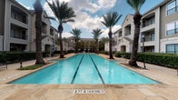 This outdoor amenity space centers around a generously sized lap pool with clearly marked lane lines, set at a uniform depth and surrounded by a broad stone-tile deck. Tall palm trees line both sides of the pool, providing a tropical aesthetic and partial shade throughout the day. The pool is framed by multi-story residential buildings constructed with warm-toned stone veneer and neutral lap siding, creating a cohesive architectural backdrop. Poolside furnishings include lounge chairs and dining-height tables with seating, offering residents a variety of relaxation and social options directly adjacent to the water. Accessible restroom facilities, including a designated men's restroom with ADA-compliant signage, are located conveniently within the pool area. A dedicated pool pump room provides on-site mechanical support for pool operations. The perimeter of the space is defined by decorative metal fencing with integrated landscape lighting, bordered by rock ground cover and mature trees that contribute to a well-maintained, resort-like environment. Walkways connect various sections of the outdoor space, ensuring easy navigation throughout the community grounds.