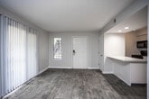 This single-floor residence presents a thoughtfully updated interior with a cohesive, contemporary aesthetic throughout. The open living area serves as the heart of the space, showcasing warm wood-look flooring, soft neutral gray walls, and clean white trim that together create a bright and welcoming ambiance. Natural light filters in through multiple windows dressed with vertical blinds, while a sliding glass door opens to the outside, further enhancing the sense of openness. A pass-through counter connects the living area to the kitchen, promoting an airy, connected flow between the two spaces. The kitchen is well-appointed with white shaker-style cabinetry, granite-look countertops, black appliances including an electric range, microwave, and dishwasher, and durable wood-look flooring that carries seamlessly from the main living area. The bedroom offers a generous, uncluttered layout with continued neutral tones and the same wood-look flooring, providing a calm and versatile space. A dedicated hallway leads to the bathroom, which features white cabinetry, a granite-look vanity top, a large mirror, and a bright vanity light fixture. The overall design prioritizes clean lines, modern finishes, and functional simplicity throughout every room.