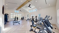 This well-appointed fitness facility spans a generously sized single floor, thoughtfully designed to accommodate a wide range of exercise disciplines. The architecture is defined by dramatic vaulted ceilings that rise to impressive heights, creating an open, airy atmosphere enhanced by clerestory windows and large multi-pane windows along the perimeter walls. Natural light floods the interior throughout the day, complemented by recessed lighting and sleek black ceiling fans that ensure comfortable air circulation.

The flooring throughout is a standout feature: light-toned hardwood laid in a classic herringbone pattern, lending an elegant yet functional aesthetic to the space. The facility is divided into distinct training zones. A large cardio area houses Matrix-brand treadmills, ellipticals, and recumbent bikes. A dedicated strength training section features cable machines, multi-functional weight stations, and adjustable benches. A functional training zone offers TRX suspension equipment, kettlebells, medicine balls, and resistance accessories neatly organized on wall-mounted racks.

A yoga and stretching area includes a large framed wall mirror, a wall-mounted television, and a decorative accent wall with patterned wallcovering in soft sage tones, adding a calm, boutique-studio quality to this section of the facility. The overall ambiance is bright, modern, and polished.