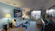 This single-floor space offers a thoughtfully designed interior that balances comfort and style. The living room serves as the centerpiece, featuring plush wall-to-wall carpeting and a warm, inviting layout. A bold teal accent wall provides a striking backdrop and sets the tone for the cohesive color palette carried throughout the space via decorative cushions and coordinating curtains.

The room is furnished with a tufted cream sofa and a complementary accent chair, arranged around a sleek glass-top coffee table with a sculptural black base. Framed artwork adorns the walls, adding a curated, personalized touch to the space. A classic table lamp introduces warm ambient lighting alongside the natural light that filters in through the sliding glass doors.

The sliding doors open to an outdoor balcony or patio, offering views of the surrounding exterior and allowing natural light to fill the room. Sheer white vertical blinds and flowing teal drapes frame the doorway elegantly. A textured ceiling adds architectural character overhead. The overall ambiance is bright, cohesive, and well-appointed, with a design sensibility that is both functional and visually appealing.
