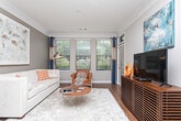 Located at 11132 Medlock Bridge Rd in Johns Creek, Georgia, this single-floor residence offers a thoughtfully designed layout with a cohesive, contemporary aesthetic carried throughout each room. The living area is an inviting and light-filled space, featuring hardwood flooring, crown molding, and a bank of large windows dressed with blue drape panels that frame views of the surrounding greenery. A plush white sofa, vibrant orange accent pillows, and a sleek floor lamp contribute to the room's modern yet comfortable character. The kitchen is equipped with rich dark-toned cabinetry, granite countertops, stainless steel appliances including an electric range and over-the-range microwave, and a tasteful tile backsplash. Pendant lighting over the sink area adds a refined finishing touch. The bedrooms are carpeted and well-proportioned, each featuring ceiling fans and windows that allow natural light to flow in. One bedroom includes a walk-in closet with built-in organization. The bathroom offers a tub-shower combination with classic white tile surround. Throughout the residence, a warm neutral color palette punctuated by orange and blue accents creates a unified, polished ambiance that is both functional and visually appealing.