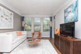 Located at 11132 Medlock Bridge Rd in Johns Creek, Georgia, this single-floor residence offers a thoughtfully designed layout with a cohesive, contemporary aesthetic carried throughout each room. The living area is an inviting and light-filled space, featuring hardwood flooring, crown molding, and a bank of large windows dressed with blue drape panels that frame views of the surrounding greenery. A plush white sofa, vibrant orange accent pillows, and a sleek floor lamp contribute to the room's modern yet comfortable character. The kitchen is equipped with rich dark-toned cabinetry, granite countertops, stainless steel appliances including an electric range and over-the-range microwave, and a tasteful tile backsplash. Pendant lighting over the sink area adds a refined finishing touch. The bedrooms are carpeted and well-proportioned, each featuring ceiling fans and windows that allow natural light to flow in. One bedroom includes a walk-in closet with built-in organization. The bathroom offers a tub-shower combination with classic white tile surround. Throughout the residence, a warm neutral color palette punctuated by orange and blue accents creates a unified, polished ambiance that is both functional and visually appealing.