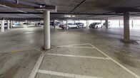 This parking facility serves a multi-story residential building and offers two distinct parking environments. The covered underground level features a concrete structure supported by cylindrical columns, with painted parking stall markings on the poured concrete floor. Overhead utilities and piping run along the low ceiling, and lighting is provided throughout. Access points within the covered level include a solid metal door marked with an illuminated EXIT sign, leading to a stairwell, as well as cinder block walls that define the perimeter of the structure. An outdoor elevator station is also available, set within a stucco-clad enclosure and equipped with standard call buttons, providing convenient vertical access between the parking level and the upper floors of the building. The open-air surface parking area sits adjacent to the building exterior, offering additional parking spaces under open sky. The surrounding building is a multi-story, stucco-finished residential structure with balconies visible along its facade. Mature trees and landscaping are present nearby, and the overall setting reflects a sunny, warm climate. The facility is clearly marked as private property, and the combination of covered and surface parking provides a practical and accessible solution for residents.