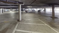 This parking facility serves a multi-story residential building and offers two distinct parking environments. The covered underground level features a concrete structure supported by cylindrical columns, with painted parking stall markings on the poured concrete floor. Overhead utilities and piping run along the low ceiling, and lighting is provided throughout. Access points within the covered level include a solid metal door marked with an illuminated EXIT sign, leading to a stairwell, as well as cinder block walls that define the perimeter of the structure. An outdoor elevator station is also available, set within a stucco-clad enclosure and equipped with standard call buttons, providing convenient vertical access between the parking level and the upper floors of the building. The open-air surface parking area sits adjacent to the building exterior, offering additional parking spaces under open sky. The surrounding building is a multi-story, stucco-finished residential structure with balconies visible along its facade. Mature trees and landscaping are present nearby, and the overall setting reflects a sunny, warm climate. The facility is clearly marked as private property, and the combination of covered and surface parking provides a practical and accessible solution for residents.