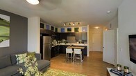 This single-floor space showcases an open-concept layout that seamlessly connects the kitchen and living areas, creating a cohesive and functional environment. The kitchen is a standout feature, designed with dark espresso cabinetry that extends to the upper walls, providing ample storage. Stainless steel appliances, including a full-size refrigerator and a black range, complement the contemporary aesthetic. A central island with a butcher block countertop doubles as a breakfast bar, accompanied by sleek white upholstered stools, making it an ideal spot for casual dining or entertaining. Track lighting overhead ensures the kitchen remains well-illuminated throughout the day. The countertops are light-toned, offering a crisp contrast to the dark cabinetry. Warm hardwood flooring runs throughout, unifying the kitchen and living areas. The living space is accented by a patterned green area rug and a casual gray sofa, adding a relaxed, inviting atmosphere. A hallway visible in the background suggests additional rooms, contributing to the overall sense of a well-organized, modern residential space.