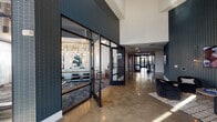 This single-floor amenity space presents a cohesive, upscale design identity from the moment you enter the lobby. Polished concrete floors and soaring ceilings define the entry corridor, which is lined with bold navy tile-clad walls and framed glass partitions that offer views into adjacent spaces. A dedicated conference and leasing area features floor-to-ceiling windows, pendant globe lighting, and a long communal bar-height table — ideal for meetings or focused work. The coworking lounge provides leather seating, wall-mounted displays, and glass-walled private rooms for quiet productivity. The resident lounge is a standout, boasting a dramatic sculptural chandelier, geometric accent walls in gold and navy, plush seating, and multiple large-screen televisions. The game room features bold mural-covered corrugated metal walls, foosball, and mounted screens for an energetic social atmosphere. A stylish banquette nook with tufted black leather seating, a marble-topped gold pedestal table, and curated artwork adds an intimate retreat. The fitness center impresses with double-height ceilings, abundant natural light from large windows, and a full array of strength and cardio equipment. The mail hall is bright and gallery-like, with geometric wall sculptures and sleek black mailboxes lining polished concrete floors.
