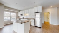 This single-floor residence showcases a contemporary design with a neutral color palette and a thoughtful layout. The open-concept kitchen and living area serves as the heart of the space, where light-toned luxury vinyl plank flooring extends throughout. The kitchen is well-equipped with a full suite of stainless steel appliances, including a French door refrigerator, electric range, built-in microwave, and dishwasher. White shaker-style cabinetry reaches toward the ceiling, offering generous storage, while a classic white subway tile backsplash adds a clean, modern touch. A central island with a warm taupe countertop provides additional prep space and casual seating. The open living area benefits from multiple windows, allowing natural light to fill the space. Bedrooms feature plush carpeting and windows with exterior views, while the primary bedroom includes a walk-in closet with wire shelving. The bathrooms are finished with white tile surrounds — one featuring a soaking tub and the other a glass-enclosed walk-in shower. A dedicated in-unit laundry closet houses a front-loading Whirlpool washer or dryer, adding everyday convenience to this well-designed residence.