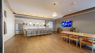 Located at 75 Tresser Boulevard in Stamford, CT, this generously sized single-floor amenity space offers a thoughtfully designed environment ideal for gathering, entertaining, and relaxing. The open layout spans a substantial footprint and is finished with warm hardwood-style flooring throughout, complemented by neutral taupe and gray wall tones that create a cohesive, contemporary aesthetic.

The living area features comfortable lounge seating, a shuffleboard table, and floor-to-ceiling windows that flood the space with natural light and frame views of the lush greenery outside. A dedicated game room includes a full-size pool table and a wall-mounted flat-screen TV, making it an inviting spot for recreation.

The dining area is anchored by a large communal table with Wishbone-style chairs, accompanied by bar-height seating along a counter, and additional high-top tables — all oriented toward another wall-mounted display. Adjacent to the dining space, a sleek wet bar with marble-style backsplash, pendant lighting, and stainless steel appliances provides a fully equipped entertaining station.

Large sliding glass doors open directly to an outdoor terrace bordered by a natural stone wall, seamlessly connecting the interior to the exterior. Recessed lighting throughout ensures a well-lit environment at any hour, while potted greenery adds warmth and life to the overall ambiance.