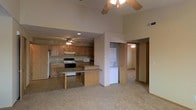 This single-floor space is defined by its open-concept layout, where the kitchen and living area flow seamlessly into one another. The kitchen is equipped with warm wood cabinetry, a white gas range, a microwave, and a refrigerator, complemented by a peninsula with a raised breakfast bar that provides both additional counter space and a casual dining option. The countertops feature a neutral laminate surface that coordinates with the cabinetry.

A standout feature of the space is the vaulted ceiling, which rises noticeably above the kitchen area, enhancing the sense of openness and volume. Ceiling fans are present in both the kitchen and living areas, supporting air circulation throughout. The living area is finished with light-toned carpeting, offering a soft contrast to the tiled kitchen floor.

A dedicated nook accommodates a stacked washer and dryer unit, providing the convenience of in-unit laundry. A hallway visible from the main living area leads to additional rooms, suggesting a functional layout with separated living and private spaces. Overall, the space offers a practical and comfortable design suited for everyday living.
