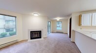 Located at 10740 NE 8th Avenue in Seattle, WA, this single-floor unit offers a well-organized layout designed for comfortable everyday living. The spacious living room serves as the heart of the home, highlighted by a white-brick wood-burning fireplace with a classic brass-trimmed glass door. Large windows along the perimeter flood the space with natural light, while a sliding glass door opens directly onto a private balcony, providing a seamless connection to the outdoors. Baseboard heating runs along the walls, and soft carpeting extends throughout the living and bedroom areas, contributing to a warm, understated ambiance. The bedroom is generously proportioned, featuring neutral walls and plush carpet, offering a quiet and versatile space. The kitchen is efficiently designed in a galley-style layout, outfitted with white cabinetry, light countertops, and white appliances including a full-size refrigerator, dishwasher, and range. Light tile flooring ties the kitchen together with a clean, practical finish. The bathroom includes a full tub-shower combination, tile flooring, a vanity with stone-look countertop, and chrome fixtures throughout. Overall, the unit presents a clean, neutral palette that provides an ideal canvas for personalization.
