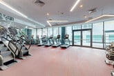 Located at 125 NE 32nd St in Miami's vibrant Edgewater neighborhood, this contemporary fitness center offers a generously sized, single-floor layout designed to accommodate a full spectrum of workout disciplines. The space is bathed in natural light, thanks to expansive floor-to-ceiling glass walls that frame views of the surrounding outdoor areas, including what appears to be a resort-style pool deck lined with lush greenery.

The main cardio zone is impressively equipped with a row of treadmills, stair climbers, and elliptical trainers, all positioned along the glass perimeter to allow exercisers to enjoy the outdoor scenery while training. A dedicated cycling studio features a fleet of stationary spin bikes arranged before a full-length mirror wall, creating an immersive group fitness environment. A functional training area rounds out the space, outfitted with a heavy bag, medicine balls, and additional strength equipment.

Throughout, the interior maintains a clean, modern aesthetic with a coffered white ceiling accented by sleek linear LED lighting, recessed fixtures, and a bold patterned carpet in warm rose and red tones. Exposed concrete columns add an architectural contrast to the polished surroundings. Double glass doors provide direct access to the outdoor terrace, seamlessly connecting indoor and outdoor fitness amenities.