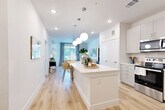 This well-designed 1-bedroom, 1-bathroom residence offers a thoughtful open-concept layout that maximizes every square foot. Light hardwood-style flooring flows seamlessly throughout, complementing the crisp white walls and creating a clean, airy aesthetic. The kitchen is a true standout, featuring white shaker cabinetry, quartz countertops, a full suite of stainless steel appliances, and a large center island with seating — perfect for dining or casual entertaining. Globe pendant lights hang above, adding a modern yet warm touch. The open layout connects the kitchen directly to the living area, which is bathed in natural light from oversized windows dressed with floor-to-ceiling drapes. The bedroom is a comfortable retreat, offering a large window with neighborhood views and direct access to the full bathroom. The bathroom impresses with dual sinks, quartz countertops, white cabinetry, and a well-lit vanity mirror. A dedicated closet provides practical storage. The hallway connects all spaces efficiently, contributing to the unit's smooth flow. Throughout, the design balances modern finishes with a light, welcoming palette that makes the space feel open and inviting.
