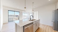 This well-appointed single-floor residence offers three bedrooms and two bathrooms within a thoughtfully designed open-concept layout. Light wood-tone flooring flows throughout the main living areas, complementing the bright, neutral palette that defines the space.

The kitchen serves as a central hub, featuring a large island with a stainless steel sink, quartz countertops, white subway tile backsplash, and stainless steel appliances including a gas range and built-in microwave. Pendant lighting with brass accents adds a modern touch above the island. The kitchen opens directly to the combined dining and living area, where expansive corner windows flood the space with natural light and provide open exterior views.

The primary bedroom features plush carpeting, a ceiling fan, and a large window with outdoor views. Additional bedrooms are also carpeted and receive generous natural light through sizable windows. The bathrooms are finished with light cabinetry, quartz countertops, and matte black hardware accents. A dedicated laundry area houses a full-size side-by-side washer and dryer. Clean white walls, panel-style doors with black hardware, and consistent finishes throughout give the space a cohesive, contemporary feel.
