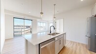This well-appointed single-floor residence offers three bedrooms and two bathrooms within a thoughtfully designed open-concept layout. Light wood-tone flooring flows throughout the main living areas, complementing the bright, neutral palette that defines the space.

The kitchen serves as a central hub, featuring a large island with a stainless steel sink, quartz countertops, white subway tile backsplash, and stainless steel appliances including a gas range and built-in microwave. Pendant lighting with brass accents adds a modern touch above the island. The kitchen opens directly to the combined dining and living area, where expansive corner windows flood the space with natural light and provide open exterior views.

The primary bedroom features plush carpeting, a ceiling fan, and a large window with outdoor views. Additional bedrooms are also carpeted and receive generous natural light through sizable windows. The bathrooms are finished with light cabinetry, quartz countertops, and matte black hardware accents. A dedicated laundry area houses a full-size side-by-side washer and dryer. Clean white walls, panel-style doors with black hardware, and consistent finishes throughout give the space a cohesive, contemporary feel.