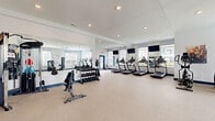 Located at 5522 Villa Rose Dr. in Powell, OH, this fitness center offers a generously sized, single-floor exercise facility designed to support a wide range of workout routines. The space is thoughtfully laid out with distinct zones for cardio, strength training, and open-floor exercise.

The cardio section lines one wall and features multiple treadmills, elliptical machines, and stationary bikes, each positioned in front of large windows that frame neighborhood views and allow abundant natural light to fill the room. Wall-mounted flat-screen televisions are installed throughout, providing entertainment during workouts. Ceiling fans help maintain comfortable air circulation across the space.

The strength training area includes a dumbbell rack with a full weight selection and a multi-functional cable machine, catering to a variety of resistance exercises. Full-length mirrors span an entire wall, visually expanding the space and allowing users to monitor their form.

A dedicated open-floor zone provides ample room for stretching, yoga, or functional training, and is stocked with stability balls in multiple sizes. Blue-trimmed glass doors connect areas of the facility, contributing to an open, airy feel. The neutral carpeted flooring, white walls, and clean architectural lines give the space a bright, modern aesthetic that is both welcoming and functional.