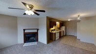 This single-floor residence offers a functional and comfortable layout designed for everyday living. The main living area is a welcoming space highlighted by a classic wood-burning fireplace with a dark wood mantle and tiled hearth, serving as a natural focal point of the room. A ceiling fan with a brass fixture overhead provides both light and air circulation throughout the carpeted living space. The living area flows seamlessly into the kitchen, which is separated by a partial wall. The kitchen is well-equipped with dark wood cabinetry offering generous storage, under-cabinet lighting, and a suite of white appliances including a range, dishwasher, and refrigerator. The kitchen also features tile flooring, providing a practical contrast to the carpeted living area. A door visible from the kitchen area suggests easy access to additional spaces or an entry point. The textured ceiling throughout adds a classic residential character to the home. Overall, the space presents a straightforward and practical layout with distinct functional zones, making it a comfortable and livable environment.