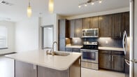 This well-appointed single-floor residence offers a thoughtfully designed layout that balances modern aesthetics with everyday functionality. The heart of the home is a contemporary open-concept kitchen, outfitted with rich dark-toned cabinetry, light quartz countertops, and a full suite of stainless steel appliances including a range, over-the-range microwave, and a bottom-freezer refrigerator. A subway tile backsplash adds a refined touch, while under-cabinet lighting highlights the clean lines of the workspace. A generously sized kitchen island with an integrated sink and pendant lighting above serves as a natural gathering point and provides additional prep space. Light-toned wood-style flooring runs throughout the kitchen and common areas, lending warmth and continuity to the space. The bedrooms are finished with plush carpeting and feature ceiling fans, large windows with blinds that invite natural light and offer outdoor views. Storage is well-considered, with a spacious walk-in closet equipped with wire shelving. The bathroom is clean and functional, featuring tile flooring, a tub-shower combination with white tile surround, a white vanity with contemporary hardware, and open built-in shelving for additional storage. Overall, the space presents a cohesive, modern design with practical amenities throughout.