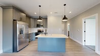 This well-appointed 2-bedroom, 1-bathroom single-floor home in Lynchburg, Virginia offers a thoughtfully designed layout with modern finishes throughout. The open-concept kitchen and living area serves as the heart of the home, anchored by a striking light blue island with a white quartz countertop, undermount sink, and pendant lighting overhead. Gray shaker-style cabinetry lines the kitchen walls, complemented by a white subway tile backsplash, white quartz counters, and a full suite of stainless steel appliances. Light wood-tone luxury flooring flows seamlessly through the main living areas, enhancing the bright, airy ambiance. A wide hallway with recessed lighting connects the living space to the private quarters. The spacious primary bedroom offers generous square footage, while the second bedroom features soft carpeting underfoot. The shared full bathroom is finished with a blue vanity, white quartz countertop, a large mirror, and a tub-shower combo. A dedicated laundry area with a stacked washer and dryer adds everyday convenience. The home's clean, neutral palette and consistent modern detailing create a cohesive and inviting atmosphere throughout.