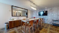 Located at 400 Walnut Street in Philadelphia's historic Old City neighborhood, this thoughtfully designed single-floor gathering space blends contemporary aesthetics with refined detailing. The centerpiece of the room is a striking floor-to-ceiling forest wallpaper mural in soft silver-blue tones, which serves as a dramatic backdrop to the wall-mounted flat-screen television and lends the space an artistic, gallery-like quality.

The flooring is a standout architectural feature — rich, dark hardwood laid in an intricate geometric pattern with gold inlay accents, adding warmth and visual depth throughout. A long bar-height marble-top table runs through the center of the room, surrounded by sculptural bentwood counter stools, making it ideal for social gatherings or collaborative events.

A built-in wet bar with warm wood cabinetry, a stainless steel sink, and a large framed mirror enhances the space's functionality and elegance. An elongated crystal chandelier anchors the main area with ambient lighting, complemented by recessed ceiling fixtures.

Natural light enters through a set of French doors at one end, which open toward an exterior corridor. Adjacent hallways provide access to elevator banks, suggesting this space functions as a shared amenity within a larger residential or mixed-use building. The overall ambiance is sophisticated, inviting, and well-appointed.