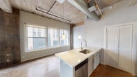 This single-floor urban loft masterfully combines industrial heritage with modern design. The open-concept living area is defined by soaring exposed concrete ceilings with visible wooden beam detailing, original ductwork, and polished concrete floors that run seamlessly throughout the space. Large multi-pane windows line the exterior wall, flooding the interior with natural light and offering views of the surrounding cityscape, while original structural columns add authentic industrial character.

The kitchen is well-appointed with gray shaker-style cabinetry, a white subway tile backsplash, stainless steel appliances including a refrigerator, electric range, and built-in microwave, and a generous center island featuring a white quartz countertop, stainless steel sink with a gooseneck faucet, and a dishwasher. Pendant lights hang above the island, adding warmth to the space.

The bedroom area benefits from the same soaring ceiling height, complete with a ceiling fan and access to closet storage. The bathroom features a white quartz double vanity with ample cabinet storage, tile-surround tub and shower combination, and polished concrete flooring consistent with the rest of the unit. Throughout, the loft strikes a compelling balance between raw, historic industrial elements and clean, contemporary upgrades.
