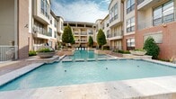 This elegantly designed outdoor amenity space is centered around a large, resort-style swimming pool that serves as the focal point of a beautifully landscaped courtyard. The pool features a classic rectangular layout with wide travertine-style coping and entry steps, reflecting the clear sky above and surrounded by carefully manicured greenery. Flanking the pool, raised planter beds and sculptural evergreen trees add vertical interest and a sense of privacy to the space.

The expansive pool deck is finished with decorative patterned stonework and is generously furnished with blue cushioned lounge chairs and shaded umbrella seating areas, creating an inviting environment for relaxation. The sundeck offers ample space for guests to enjoy the outdoors in comfort and style.

Enclosing the courtyard on multiple sides is a multi-story building constructed with warm red brick and accented with large-pane windows, balconies with metal railings, and architectural curved rooflines that lend the structure a refined, contemporary character. Gated access points connect the pool area to interior corridors, ensuring security and convenience. Along the building's perimeter, well-maintained garden beds with ferns, tropical plantings, and decorative stone borders line the paved walkways, adding lush, natural texture throughout the space.