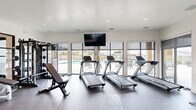 Located in Spanish Fork, Utah, this modern fitness center offers a comprehensive workout facility designed to meet a wide range of exercise needs. The single-level space is thoughtfully laid out, maximizing its generous footprint to accommodate both cardio and strength training zones.

Large, floor-to-ceiling windows line one side of the room, flooding the interior with natural light and providing direct views of an outdoor pool and patio area. Roller shades offer adjustable light control throughout the day. A wall-mounted flat-screen television is centrally positioned, visible from the cardio area.

The cardio section features multiple treadmills and an upright stationary bike, arranged to take full advantage of the poolside views. On the strength side, the space is equipped with a multi-station cable machine, adjustable weight benches, a dumbbell rack with a full range of weights, medicine ball storage, and additional selectorized machines. Full-length mirrors are mounted along one wall, enhancing the sense of openness and functionality.

A glass partition wall separates a secondary strength training area, providing a semi-private space for focused workouts. Durable dark flooring runs throughout, and recessed lighting keeps the environment bright and welcoming. Double glass doors open directly to the outdoor amenity area, seamlessly connecting indoor and outdoor fitness experiences.