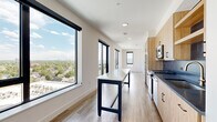 Located at 1300 40th St in Denver, CO, this modern one-bedroom, one-bathroom unit offers a thoughtfully designed open layout with contemporary finishes throughout. Large black-framed windows flood every room with natural light and frame impressive elevated views of the Denver skyline, surrounding neighborhoods, and distant mountain ranges.

The kitchen is a standout feature, equipped with light wood cabinetry, dark quartz countertops, a bold slate-blue subway tile backsplash, stainless steel appliances including a gas range and microwave, a gold-toned faucet, and a stainless steel island that doubles as a prep and dining surface. The living area flows naturally from the kitchen, benefiting from the same abundant natural light and access to an outdoor balcony.

The bedroom is carpeted for comfort, features large black-framed windows with sweeping city views, and connects directly to a dedicated closet with built-in shelving and wall hooks. Light wood-look flooring extends through the main living areas and hallways, adding warmth to the clean, white interior palette.

The bathroom is finished with a floating wood vanity, dark quartz countertop, undermount sink, gold faucet hardware, black fixtures, a modern multi-bulb pendant light fixture, a full-length mirror, and a floor-to-ceiling black-tiled glass-enclosed walk-in shower. The entrance completes this well-appointed unit with a welcoming transitional space.