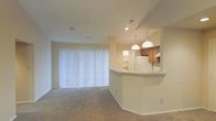 This single-floor residence showcases an open-concept design that seamlessly connects the living area and kitchen. The carpeted living space is generously sized, featuring neutral beige walls and a large sliding glass door dressed with vertical blinds, allowing for ample natural light and potential outdoor access. The kitchen is defined by a curved, raised breakfast bar that serves as a natural divider between the two spaces, offering both functional counter seating and an aesthetic architectural detail. Warm-toned wooden cabinetry lines the kitchen walls, complemented by white appliances including a refrigerator. Pendant lights hang above the breakfast bar, providing focused task lighting while adding a decorative element to the space. A built-in shelving nook is visible near the kitchen, offering additional display or storage opportunities. Recessed lighting in the kitchen area, combined with the pendant fixtures, ensures the space is well-lit. The overall color palette remains consistent throughout, with light beige tones creating a cohesive and neutral ambiance across the living and kitchen areas.