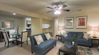 This single-floor residence offers a thoughtfully designed open-concept layout that seamlessly connects the main living spaces. The living room serves as the heart of the home, featuring warm neutral-toned walls, light carpeting, and a classic ceiling fan with integrated lighting that adds both function and a finishing touch to the space. The room comfortably accommodates a full seating arrangement, with ample space for relaxing or entertaining.

Adjacent to the living room, a breakfast bar with bar-height seating provides a casual dining option and acts as a natural divider between the living area and the kitchen. The kitchen is visible from the main living space and features dark cabinetry, a full-size stove, and a refrigerator, offering all the essentials for home cooking.

A dedicated dining area is also present, positioned near the kitchen for easy access during mealtimes. Decorative framed artwork on the walls and tasteful furnishings throughout contribute to a cohesive, welcoming ambiance. The layout is practical and efficient, making excellent use of the available floor space while maintaining an open, airy feel across all areas of the home.