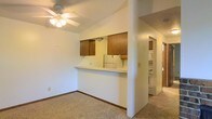 This single-floor residence offers a practical and comfortable layout designed for everyday living. The living area features wall-to-wall carpeting that adds warmth underfoot, and a ceiling fan with a light fixture provides both air circulation and ambient lighting overhead. A raised breakfast bar serves as the dividing element between the living space and the kitchen, creating an open, connected feel while maintaining a defined sense of each area. The kitchen is equipped with warm-toned wooden cabinetry and white appliances, including a refrigerator, offering a classic and functional workspace. A range hood above the cooking area adds to the kitchen's practicality. Just off the main living area, a hallway leads to additional rooms, with a glimpse of what appears to be a dedicated laundry area, complete with stacked or side-by-side units — a convenient in-unit feature. The neutral color palette throughout, with off-white walls and light carpet tones, creates a clean and versatile backdrop. The open sightlines across the space contribute to an airy, welcoming ambiance suitable for a variety of living arrangements.