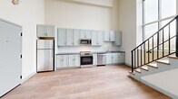 Located at 160 Seabury St in Fall River, MA, this two-bedroom, one-bathroom unit spans two floors and showcases a thoughtful blend of architectural character and modern finishes. The main floor comprises the larger portion of the living space and is anchored by a well-appointed kitchen featuring light gray shaker-style cabinetry extending to impressive heights, dark granite countertops, a white subway tile backsplash, and a full suite of stainless steel appliances including a range, built-in microwave, dishwasher, and refrigerator. Light hardwood flooring runs throughout both levels, lending warmth and continuity to the space. A staircase with a metal railing connects the two floors, adding an industrial-modern touch. The upper level hosts the bedrooms, including a primary bedroom fitted with a double closet. A striking arched window with a black metal frame and interior railing is a defining architectural feature, flooding the space with natural light and offering views of the surrounding neighborhood. The full bathroom includes a tub and shower combination with glass sliding doors and granite-topped vanity cabinetry. Neutral warm tones throughout create a clean, inviting ambiance.