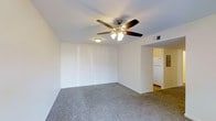 This single-floor residence at Willowbrook Apartments offers a straightforward and functional layout designed for comfortable everyday living. The main living area is finished with wall-to-wall carpeting in a neutral gray tone, providing a soft underfoot feel that complements the clean, white walls throughout the space. A five-blade ceiling fan with an integrated light kit is centrally mounted overhead, offering both air circulation and ambient lighting. An additional wall-mounted light fixture provides supplemental illumination. The walls feature a subtle textured finish, adding a modest design detail to the otherwise clean interior. A standard HVAC vent is visible near the ceiling, indicating a climate control system is in place. An open passageway connects the living area to the kitchen, which is glimpsed in the background and appears to include a full-size refrigerator and transitions to a hard-surface floor. The overall ambiance is simple and move-in ready, with a neutral color palette throughout that offers flexibility for a variety of furnishing styles. The single-floor design ensures easy accessibility and convenient navigation throughout the home.