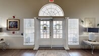 Silver Elm is a thoughtfully designed senior living facility spanning a generous single floor, offering a welcoming and refined environment throughout. The entrance makes an immediate impression with a set of classic white French doors framed by decorative columns and a dramatic arched transom window that floods the foyer with natural light. Flanking sidelights with plantation shutters complete the elegant entry aesthetic.

The spacious lobby and reception area features a curved teal front desk, warm wood-look flooring, recessed lighting, and a decorative aquarium that adds a lively focal point. The long, open corridor extends the facility's reach, lined with tasteful wall décor, pendant lighting, and blue accent doors that provide visual interest.

The common living areas are generously proportioned with soaring vaulted ceilings, arched windows, and an abundance of natural light. A comfortable lounge area includes a large sectional sofa, chandelier, and coffee table arranged for relaxed gatherings. A fireplace with a classic white mantle anchors another seating area, complemented by elegant accent chairs and refined artwork.

A dedicated dining area features multiple tables with upholstered seating beneath a chandelier, serving as a communal hub. Throughout, warm neutral tones, crown molding, wainscoting, and wood-look flooring create a cohesive, hospitable ambiance.