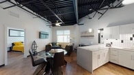 This single-floor urban loft blends industrial character with modern comfort in a striking open-concept layout. The defining architectural feature is the soaring exposed black steel beam ceiling, which lends dramatic height and an authentic loft aesthetic throughout the main living area. Warm hardwood flooring runs continuously underfoot, connecting each zone seamlessly.

The kitchen is well-appointed with white shaker cabinetry, bright quartz countertops, a classic white-and-black tile backsplash, and a full suite of stainless steel appliances including a French door refrigerator, range, microwave, and dishwasher. A large island provides additional prep and dining space.

The open dining area accommodates a round glass table, while the living room offers generous seating with multiple dark leather sofas, a wall-mounted flat-screen TV, and large windows that flood the space with natural light. A ceiling fan overhead adds both function and style.

The bedroom is a calm retreat featuring dual windows, tall ceilings with exposed beams, ample closet space, and warm hardwood floors. The bathroom is finished with bold black-and-white square tile accents, a glass-enclosed walk-in shower with floor-to-ceiling tile surround, and hardwood flooring that carries through cohesively.