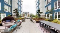 This outdoor courtyard is set within a contemporary multi-story building complex characterized by bold blue and white facades with large windows. The expansive tiled terrace serves as a central gathering space, thoughtfully designed to accommodate a variety of social settings. Large-format concrete pavers create a clean, cohesive foundation throughout the courtyard, lending a polished, modern aesthetic.

The space is divided into distinct functional zones. A dining area features a generous long table with seating for multiple guests, complemented by smaller round bistro-style tables and chairs positioned on patterned area rugs. Toward one end, striking marble-accented outdoor kitchen or grill stations anchor the space near large glass doors that connect the courtyard to an interior amenity room.

A relaxed lounge area offers woven pod chairs with canopied day beds and rattan seating adorned with colorful cushions, creating a comfortable retreat. Raised planting beds filled with ornamental grasses, ferns, manicured pine trees, and ground cover add natural texture and greenery, softening the architectural surroundings. The overall ambiance balances urban sophistication with an inviting, resort-like atmosphere suitable for casual lounging or communal dining.